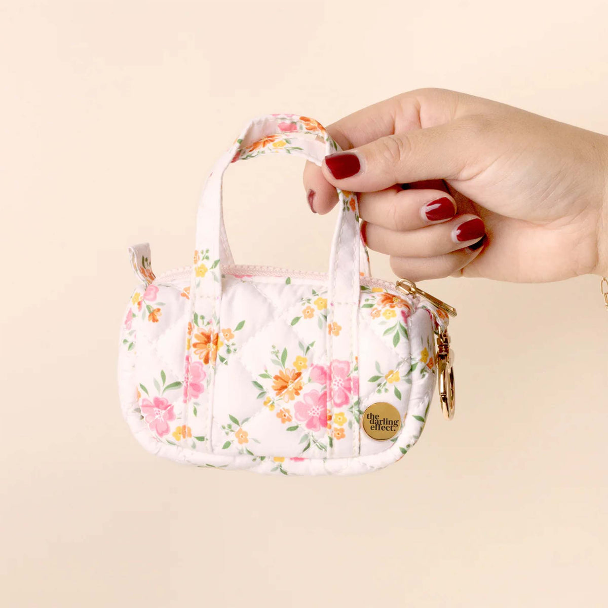 Hand holding the white floral mini duffle keychain, displaying the front pattern, top handles, and gold charm logo.