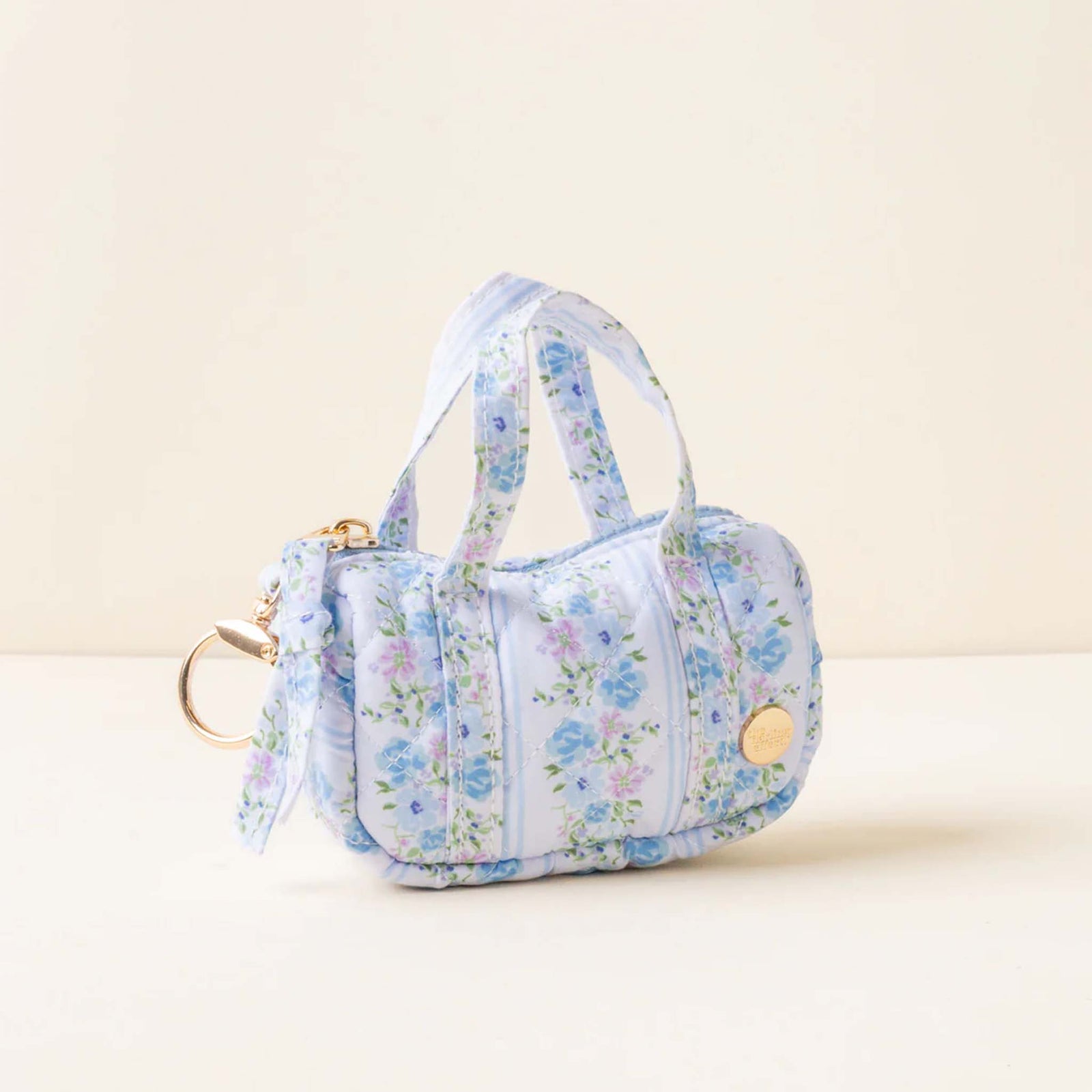 Side view of a pastel blue mini duffle keychain with blue and lavender floral print and gold keyring.