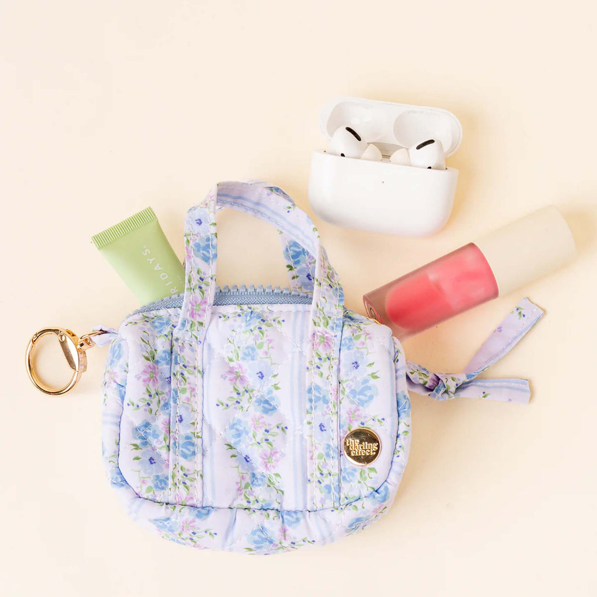 Mini floral keychain bag with lip gloss, hand cream, and AirPods arranged beside it to show scale.