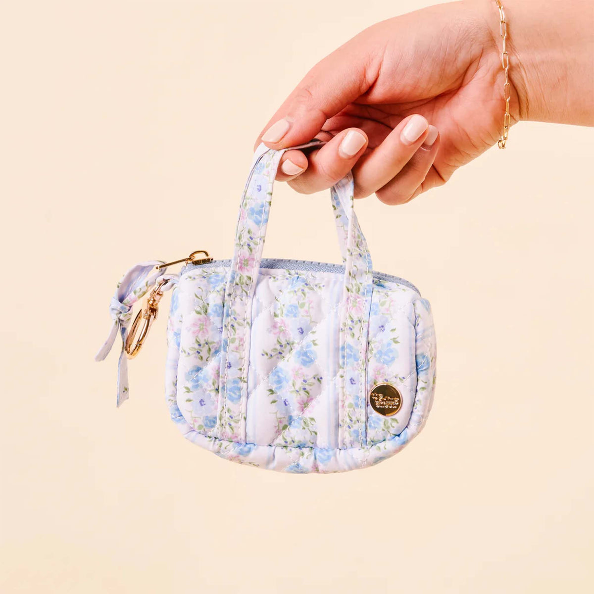 Hand holding the pastel blue floral mini duffle keychain, highlighting its quilted design and gold logo charm.