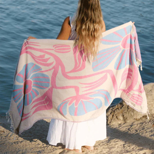 Woman holding a pink and blue floral patterned Sand Cloud towel by the water