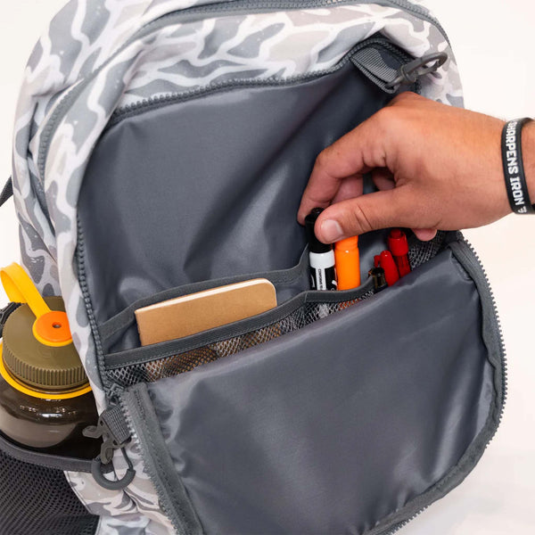 Gray camouflage backpack with a hand reaching in to organize items.