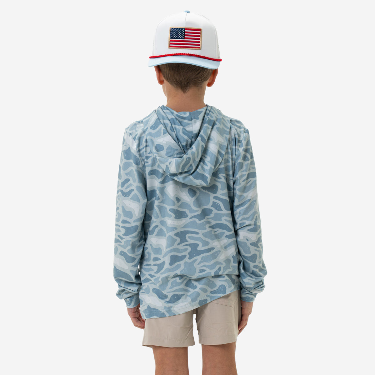 Child wearing a camouflage long-sleeve shirt and beige shorts with an American flag cap on a white background