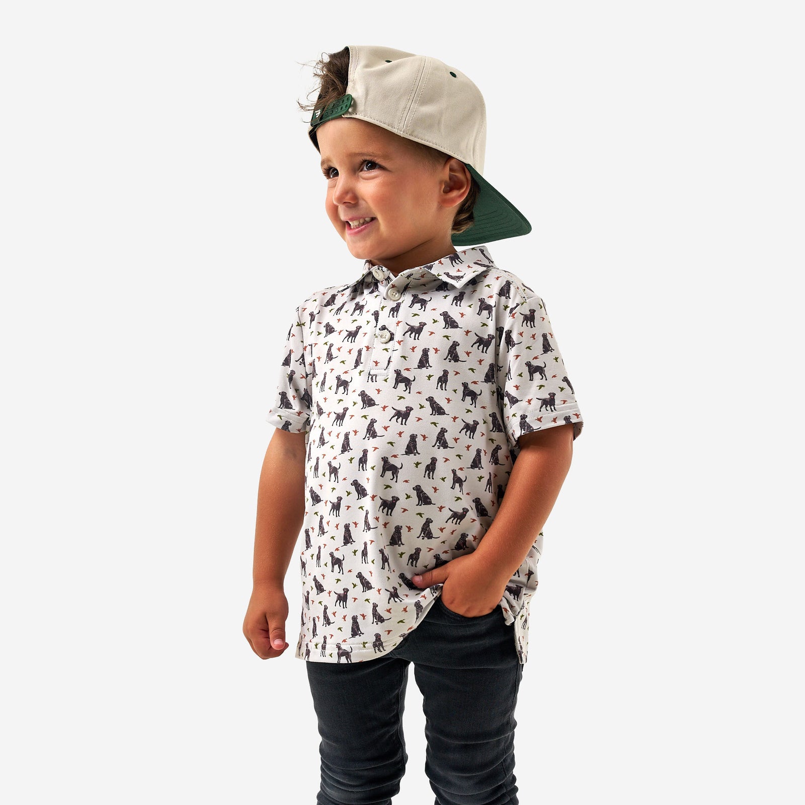 Child wearing a Burlebo polo shirt with all over pattern of ducks and dogs and a backwards cap on a white background.
