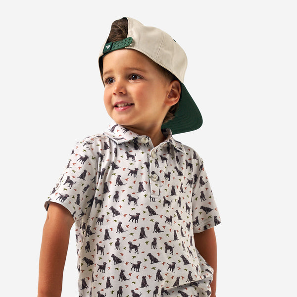 Child wearing a Burlebo polo shirt with all over pattern of ducks and dogs and a backwards cap on a white background.