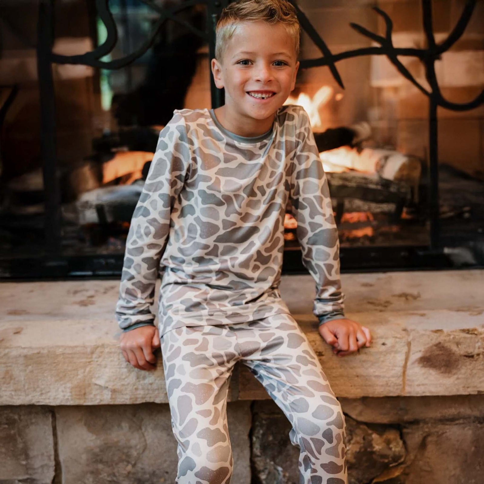 A young boy wearing gray and brown camouflage pajamas, sitting on a stone ledge in front of a fireplace, smiling with his legs crossed.