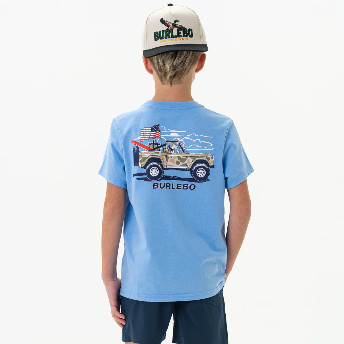 Burlebo USA Cruiser Youth Short Sleeve T-shirt, color light blue, youth sizes S-L, 50% cotton, 50% polyester, pre-shrunk.
A young boy is shown from the back wearing a light blue short-sleeve T-shirt and dark navy shorts. The shirt features a graphic of a tan camouflage off-road vehicle with an American flag and a red Burlebo flag flying from the back. A dog is illustrated sitting in the driver's seat, and white clouds stretch across the sky in the background. The word "BURLEBO" is printed below the graphic.